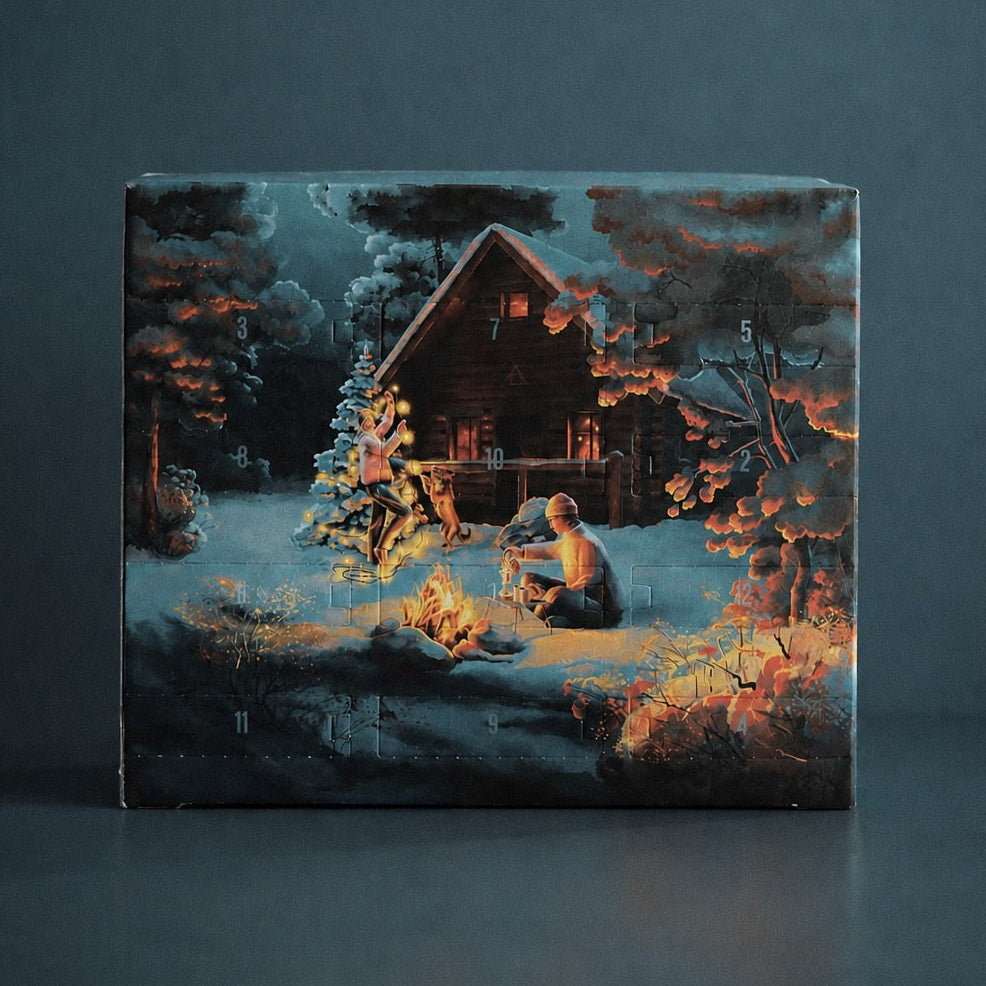 A rectangular decorative box or canvas featuring a painted winter night scene with a cabin, snowy ground, trees, and a person by a warm fire, photographed against a dark blue-gray studio background.